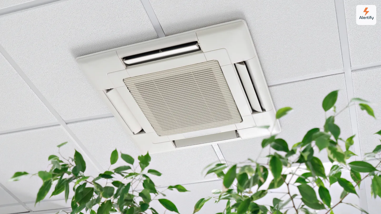 Uma unidade de ar condicionado montada no teto, embutida num teto de azulejos, com as folhas de uma planta de interior em primeiro plano.