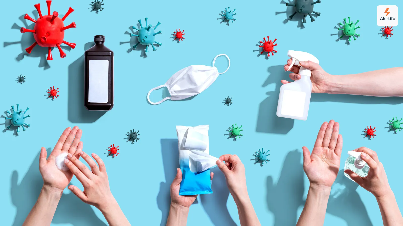 Vista de cima de modelos de vírus, uma máscara, um spray desinfetante e mãos a usar produtos de higiene
