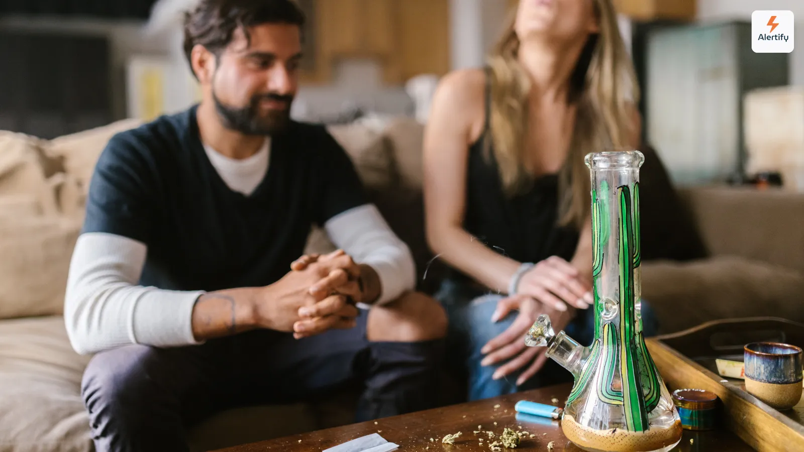 Dois hóspedes sentados num sofá na sala de estar de um alojamento, com um bong de vidro, um isqueiro e um moinho em cima da mesa de centro à sua frente.